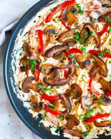 Frying pan from above with fettuccini pasta in a white-orange creamy sauce with sliced crispy mushrooms, red bell pepper sticks and freshly chopped parsley.
