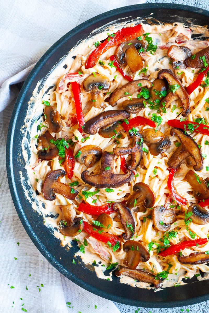 Frying pan from above with fettuccini pasta in a white-orange creamy sauce with sliced crispy mushrooms, red bell pepper sticks and freshly chopped parsley.