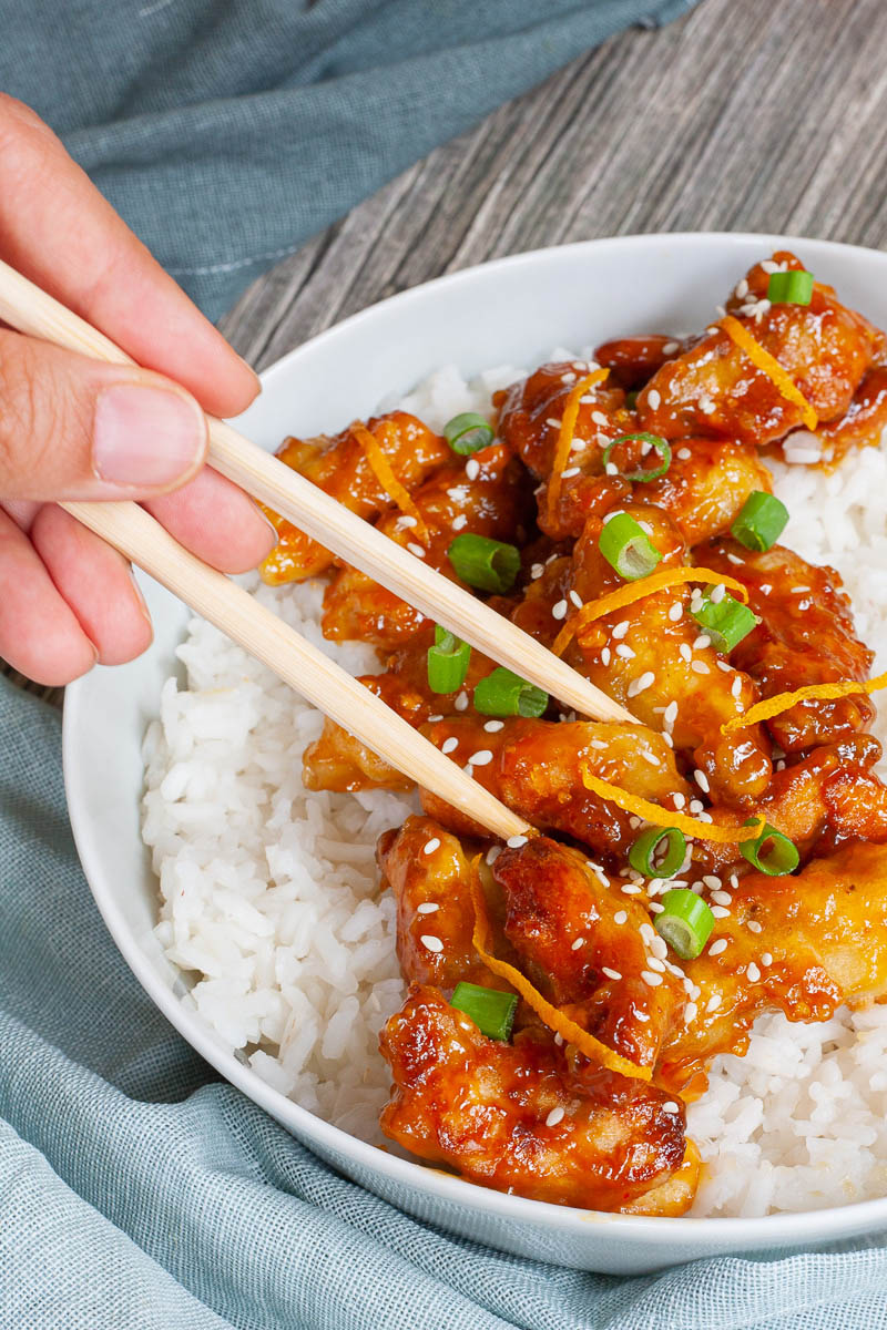 A large white bowl with white rice and sticky brown battered strips on top sprinkled with sesame seeds, orange peels and chopped spring onion. A hand is holding chopsticks and taking on strip.