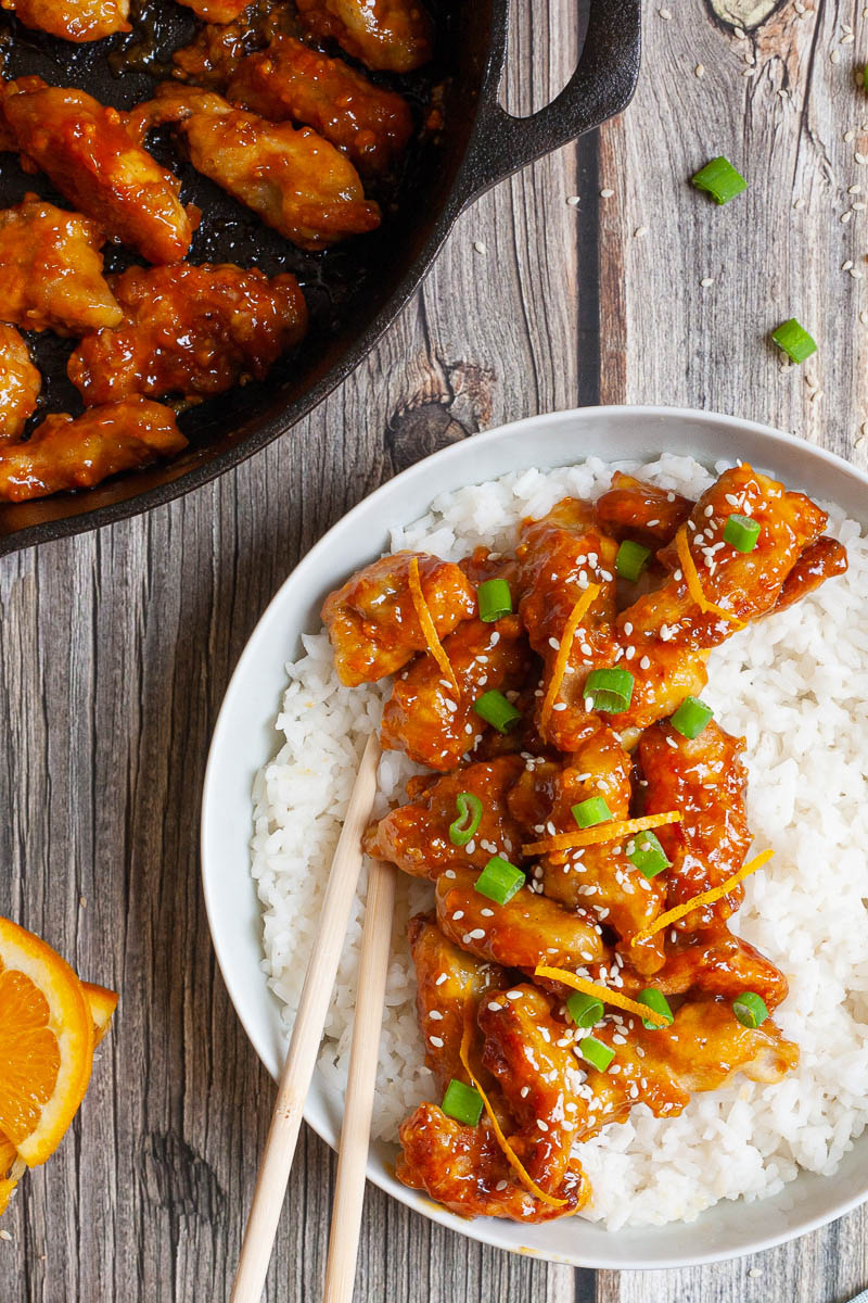 A large white bowl from above with white rice and sticky brown battered strips on top sprinkled with sesame seeds, orange peels and chopped spring onion. A black cast-iron skillet with the remaining strips is in the corner.