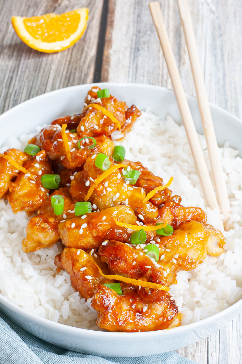 A large white bowl with white rice and sticky brown battered strips on top sprinkled with sesame seeds, orange peels and chopped spring onion