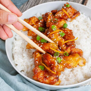 A large white bowl with white rice and sticky brown battered strips on top sprinkled with sesame seeds, orange peels and chopped spring onion. A hand is holding chopsticks and taking on strip.