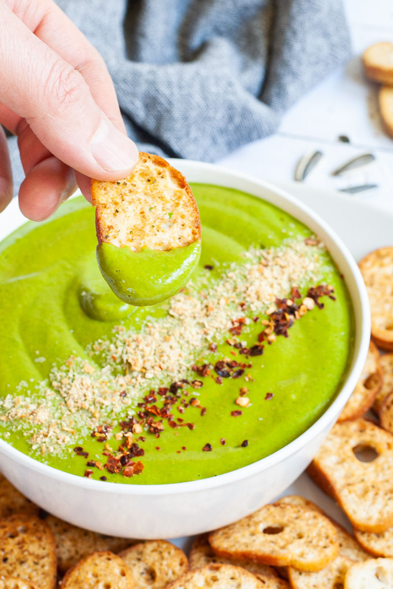 A white bowl with green sauce sprinkled with yellow flakes and red chili flakes. A hand is holding a piece of toasted bread to dip it in. More small toasted bread pieces are around the bowl.