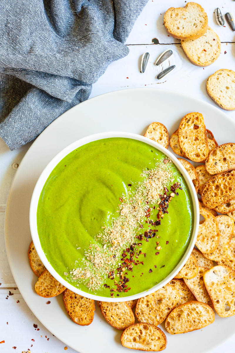 A white bowl from above with green sauce sprinkled with yellow flakes and red chili flakes. More small toasted bread pieces are around the bowl.