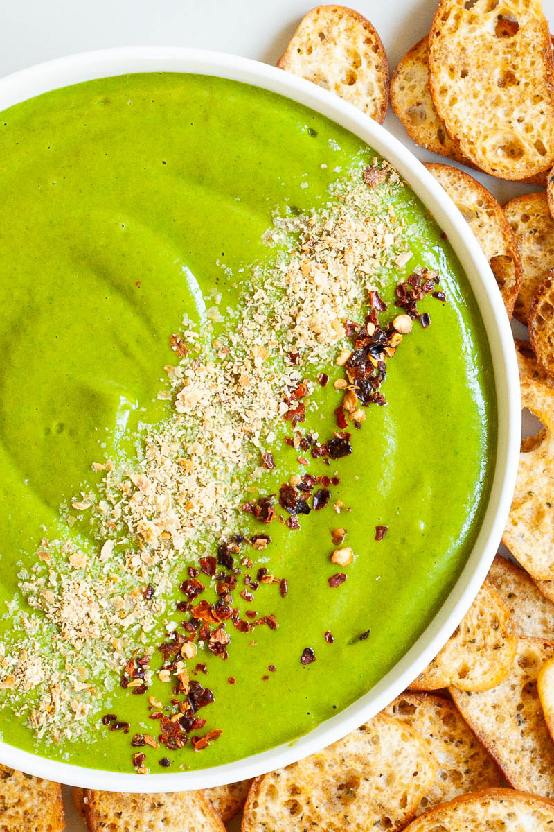 A white bowl from above with green sauce sprinkled with yellow flakes and red chili flakes. More small toasted bread pieces are around the bowl.