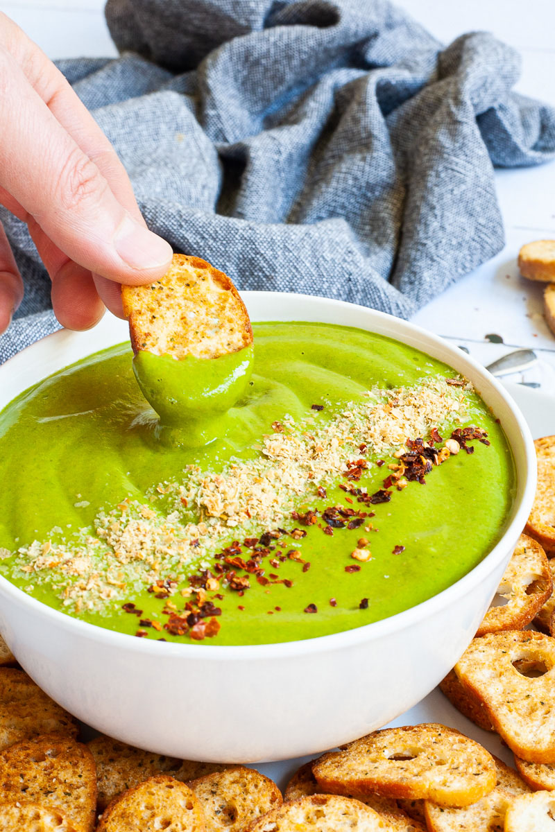 A white bowl with green sauce sprinkled with yellow flakes and red chili flakes. A hand is holding a piece of toasted bread to dip it in. More small toasted bread pieces are around the bowl.