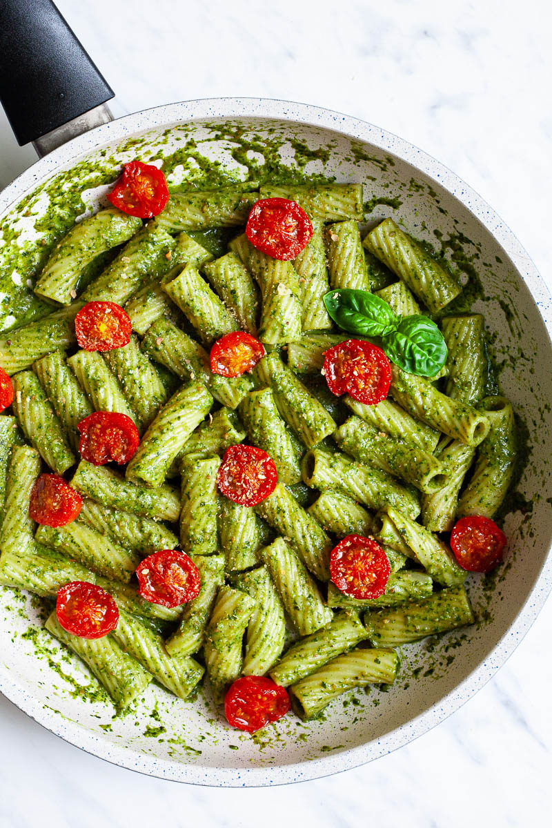 White frying pan with tortiglioni pasta covered in green paste, wilted half cherry tomatoes are on top scattered.