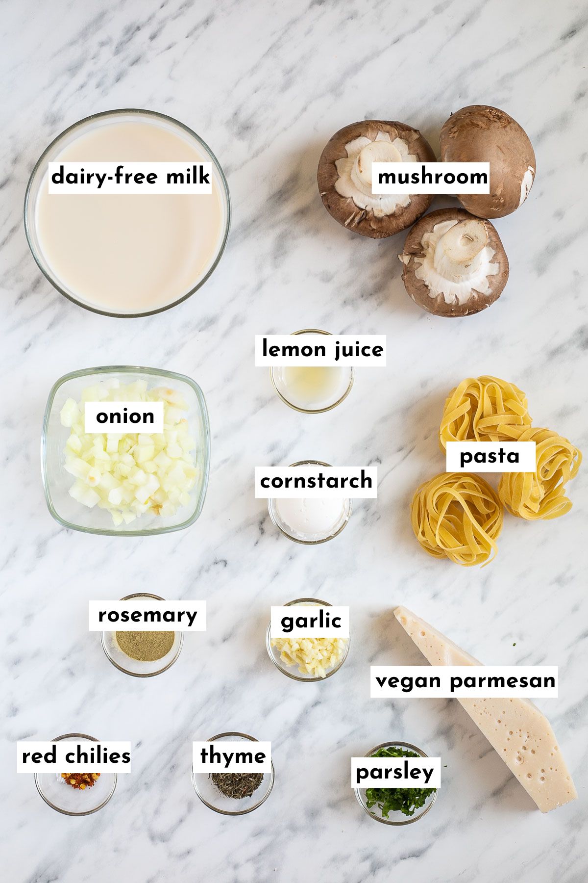 The ingredients of vegan mushroom pasta is in small glass bowls like milk, chopped onion, garlic, rosemary, lemon juice, starch, parsley, thyme, parmesan piece, dry tagliatelle. 