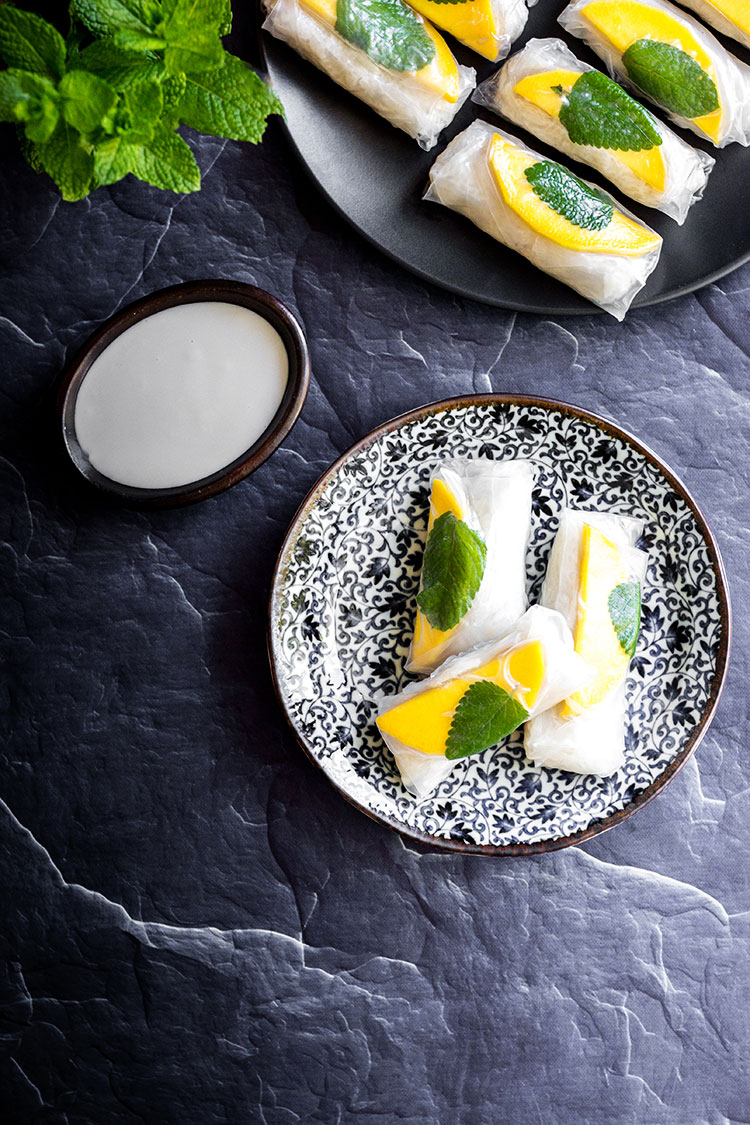 Small plate with black pattern on a grey stone surface with 3 transparent rolls where you can see white rice, mango slices and mint leaves