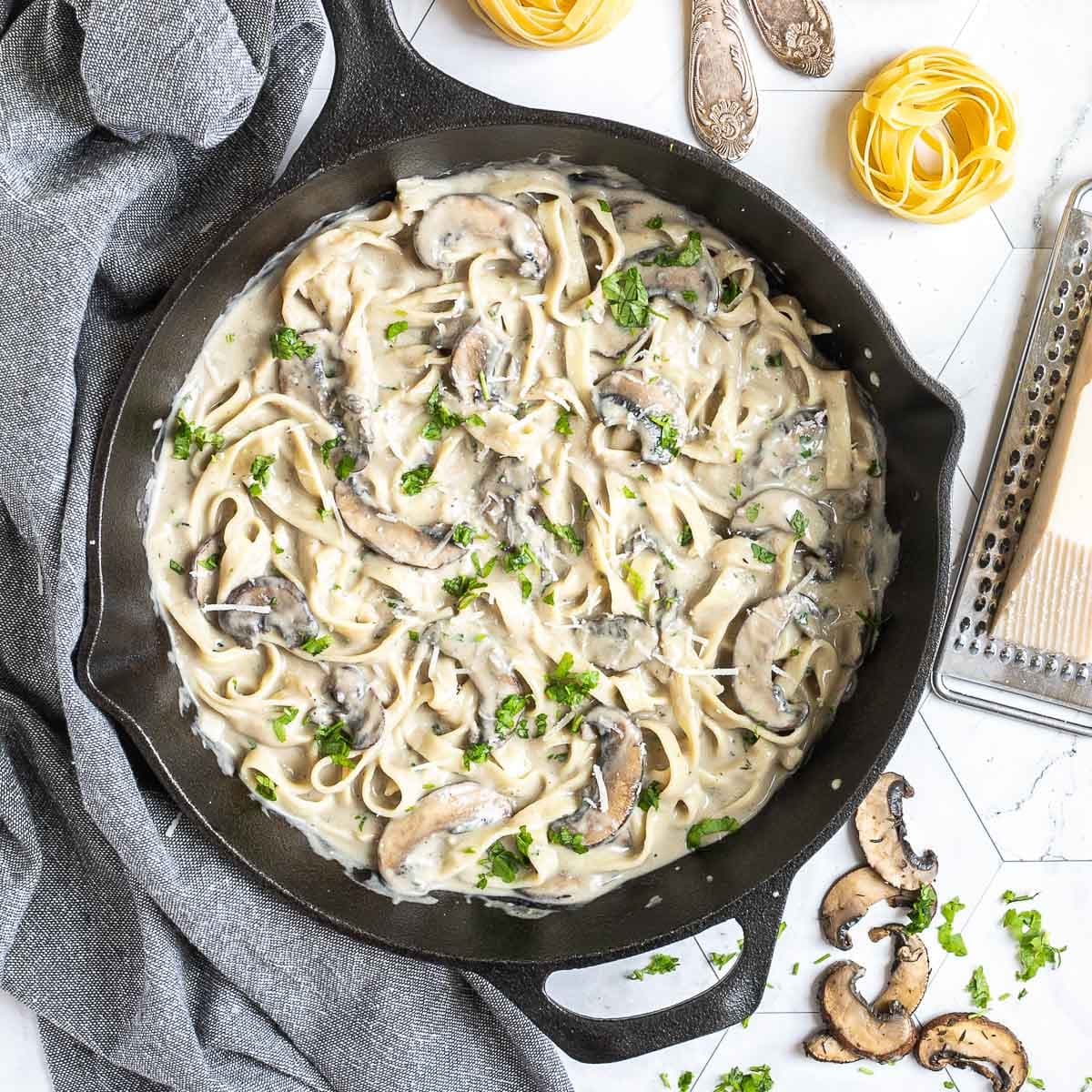 Cast iron skillet with tagliatelle in a creamy light brown sauce with sliced mushrooms and freshly chopped green herbs. Dry tagliatelle and a cheese grater with a stick of parmesan is next to it.