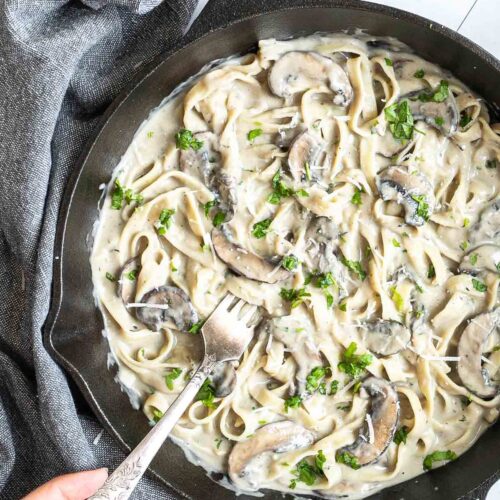 Cast iron skillet with tagliatelle in a creamy light brown sauce with sliced mushrooms and freshly chopped green herbs. A hand is holding a fork to the middle.