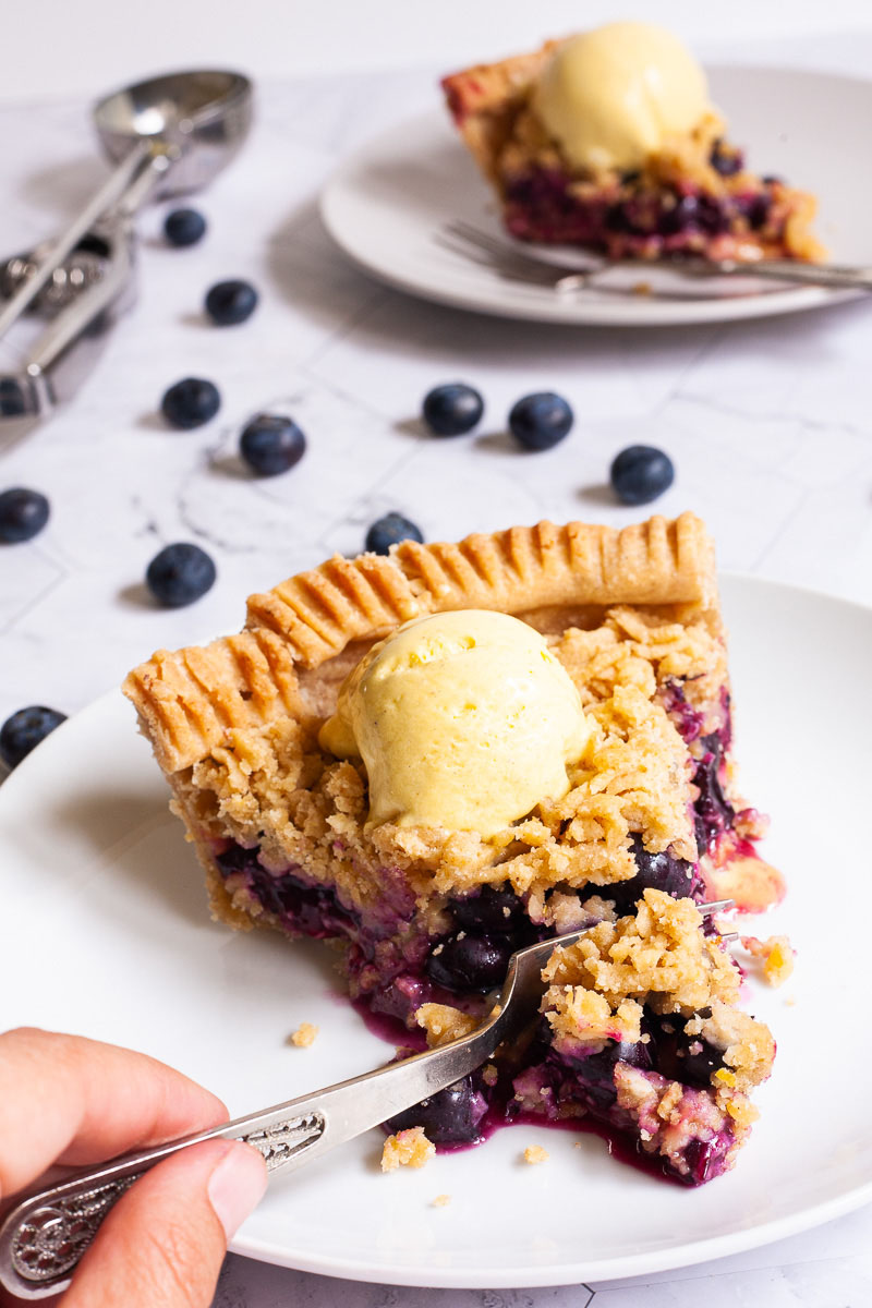 One slice of blueberry pie on a white plate with vibrant purple berries, crumble topping and a scoop of yellow ice cream. Another plate with a slice is in the background. Fresh blueberries are scattered around.
