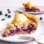 One slice of blueberry pie on a white plate with vibrant purple berries, crumble topping and a scoop of yellow ice cream. Another plate with a slice is in the background. Fresh blueberries are scattered around.