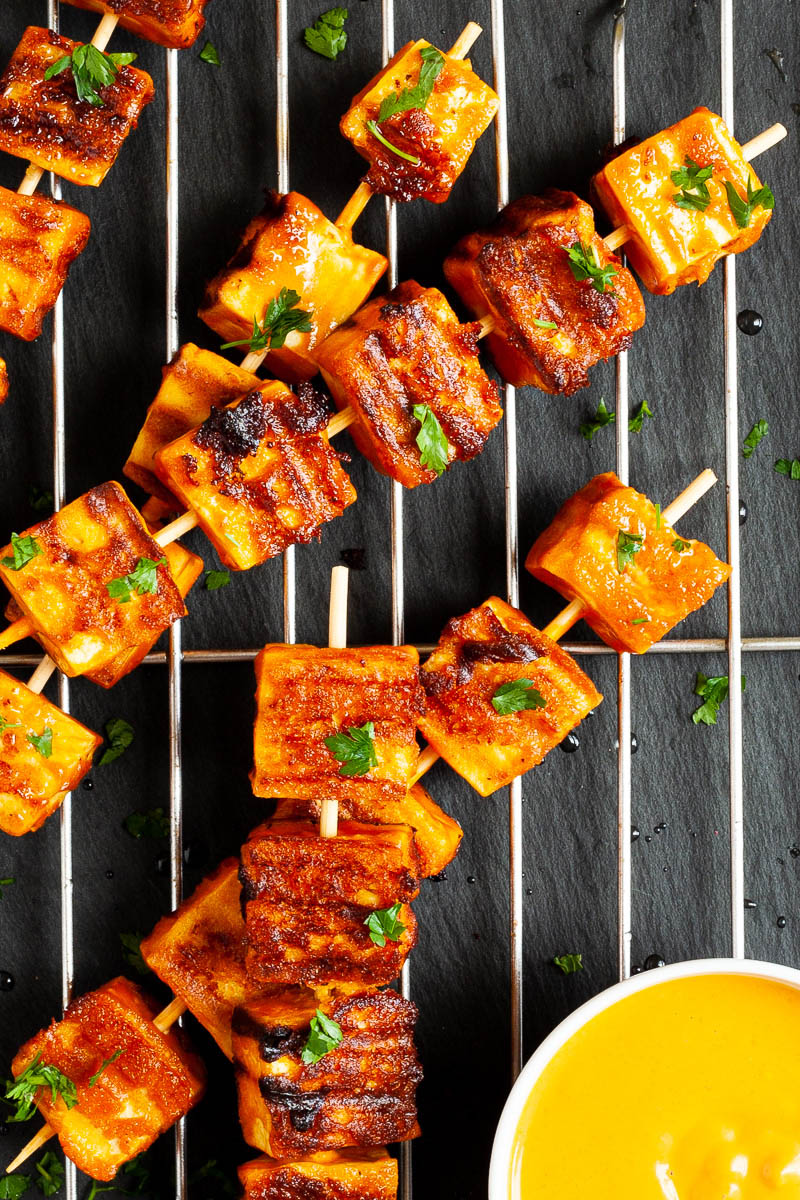 Yellow-brown tofu cubes on wooden skewers are criss cross over each other on a grilling rack placed on a black board. A small white bowl is next to them with a yellow dipping sauce.
