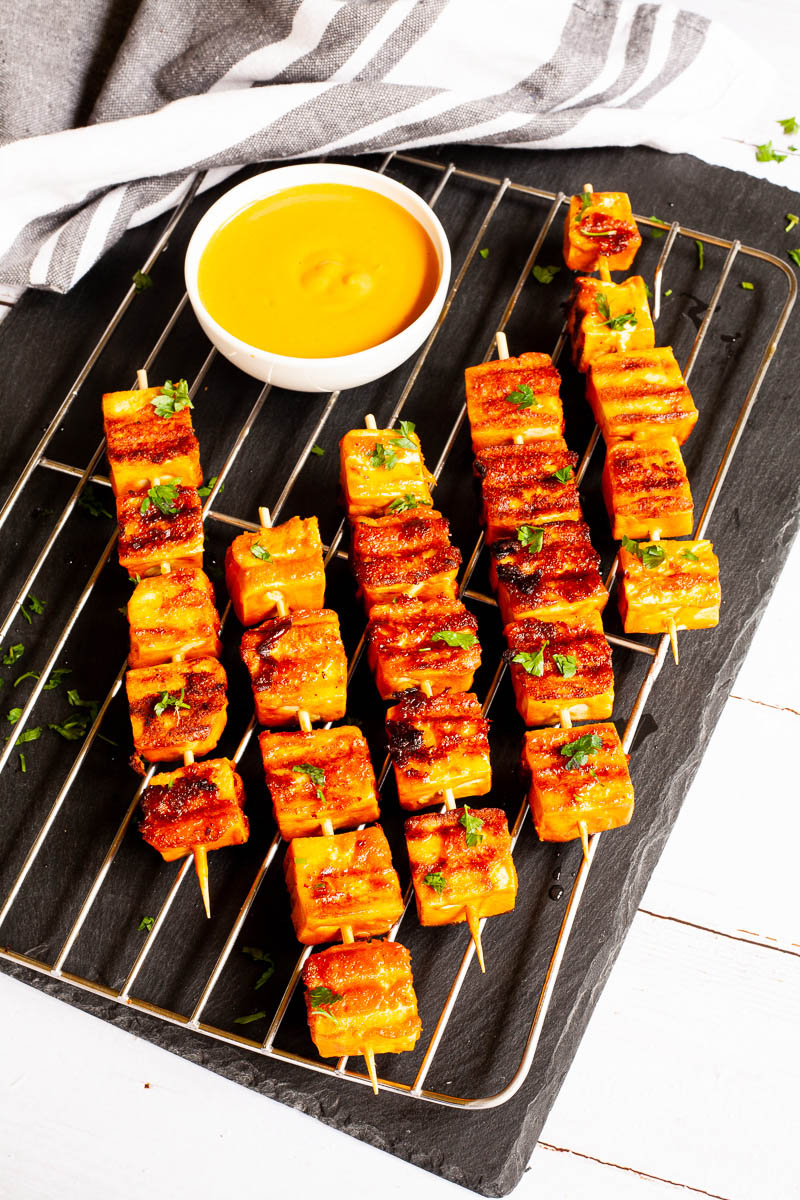 Yellow-brown tofu cubes on wooden skewers are criss cross over each other on a grilling rack placed on a black board. A small white bowl is next to them with a yellow dipping sauce.