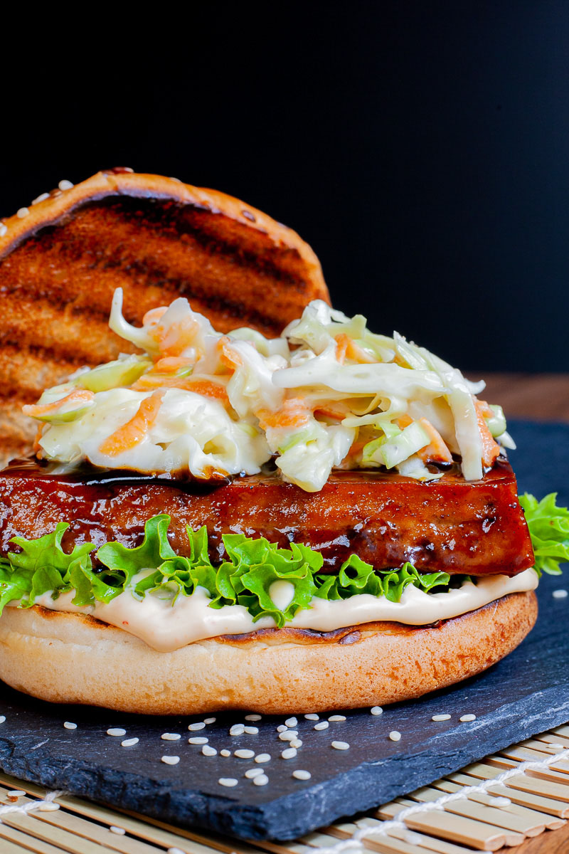 A burger bun up close with white sauce, green ruffled lettuce, dark glazed tofu slice, shredded cabbage and carrot in white sauce. The top bun is placed to the side with charcoal marks.