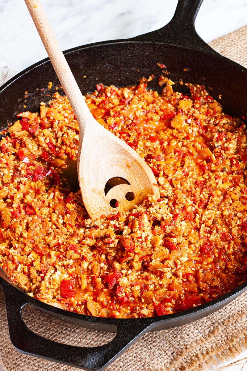 A black cast iron skillet from above with yellow brown pieces in a red chunky sauce. There is a wooden spatula with a smiley face on it placed across the sides.