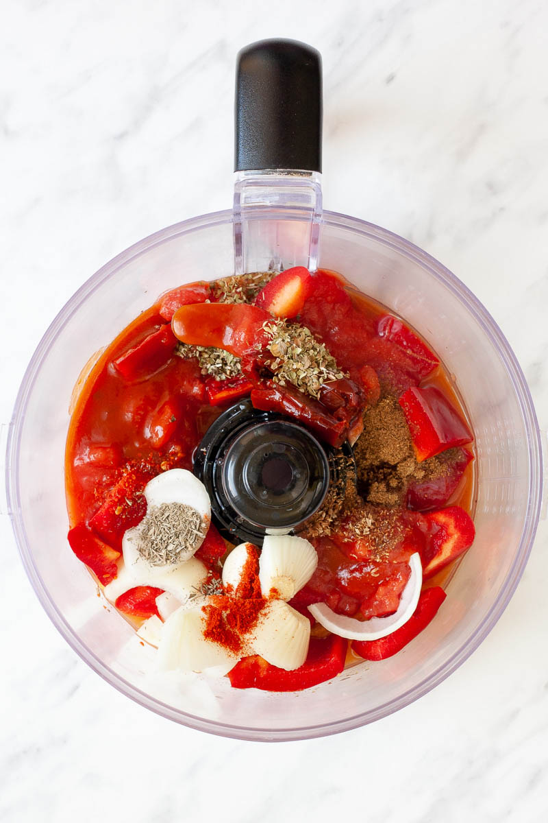 A food processor from above showing a red bell pepper chunks, tomato chunks, red tomato sauce, white onion, lots of brown and dried green herbs