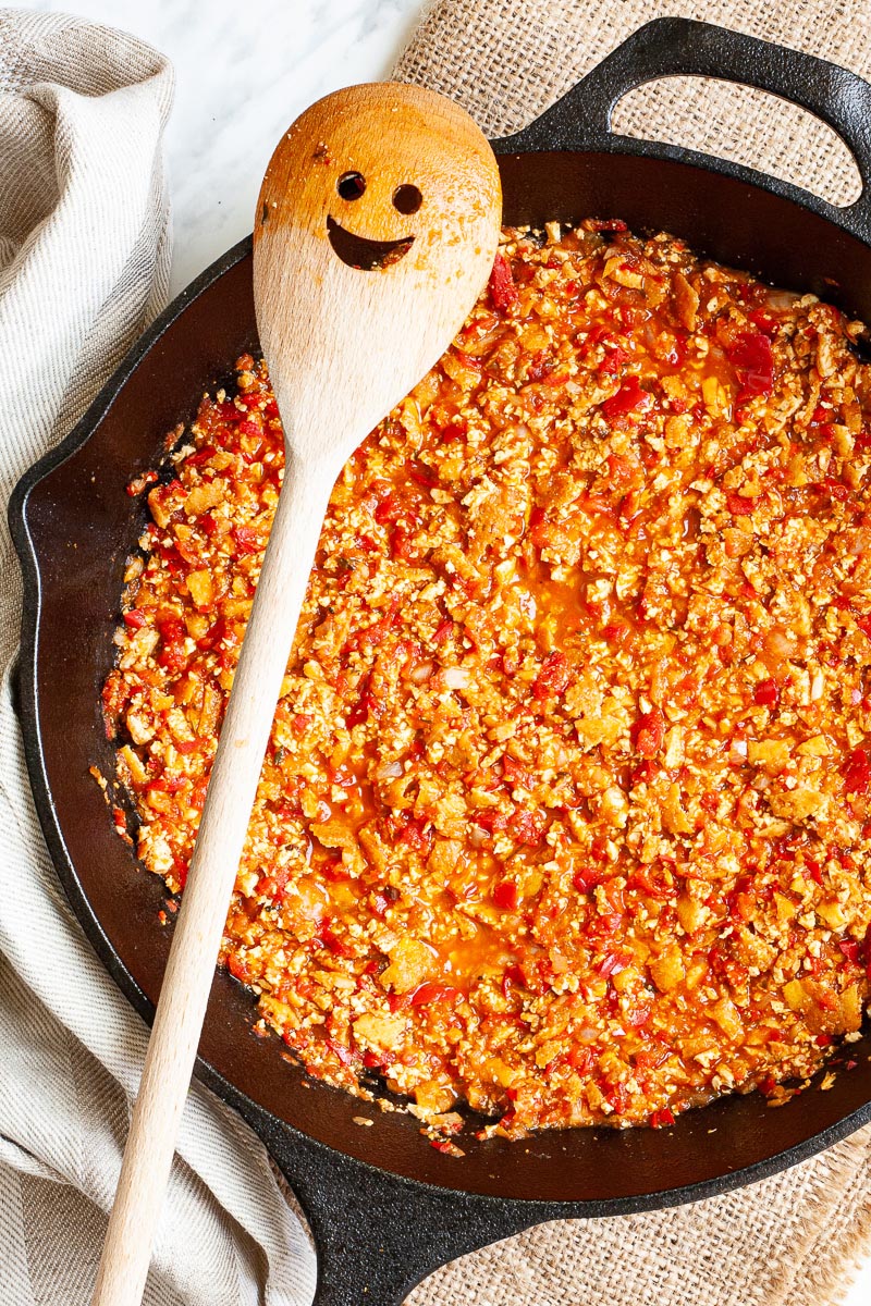 A black cast iron skillet from above with yellow brown pieces in a red chunky sauce. There is a wooden spatula with a smiley face on it placed across the sides.