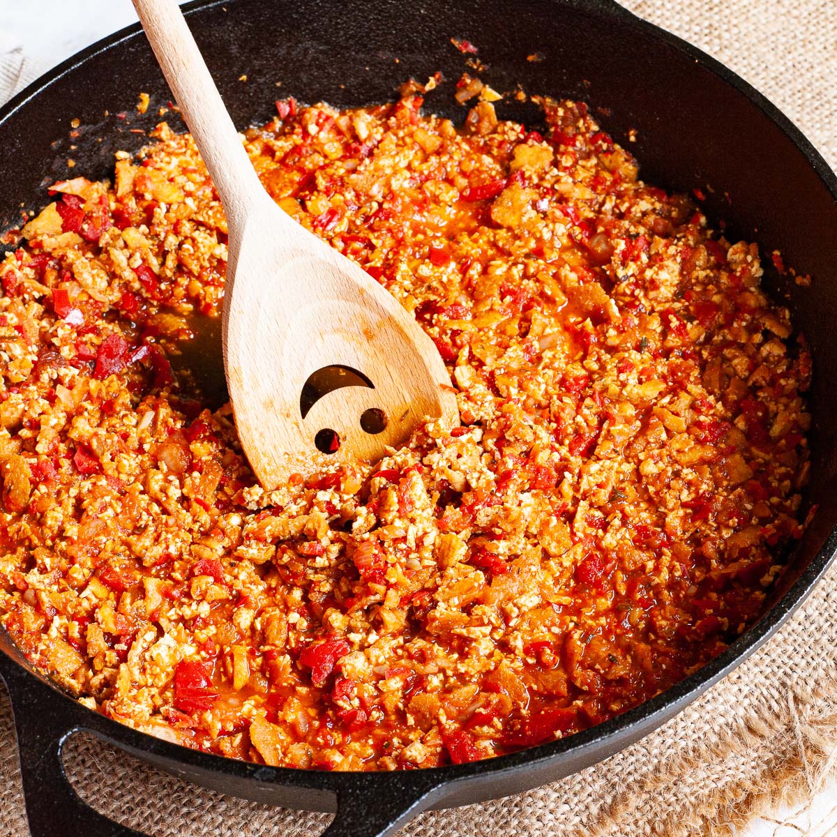 A black cast iron skillet from above with yellow brown pieces in a red chunky sauce. There is a wooden spatula with a smiley face is stirring the middle.