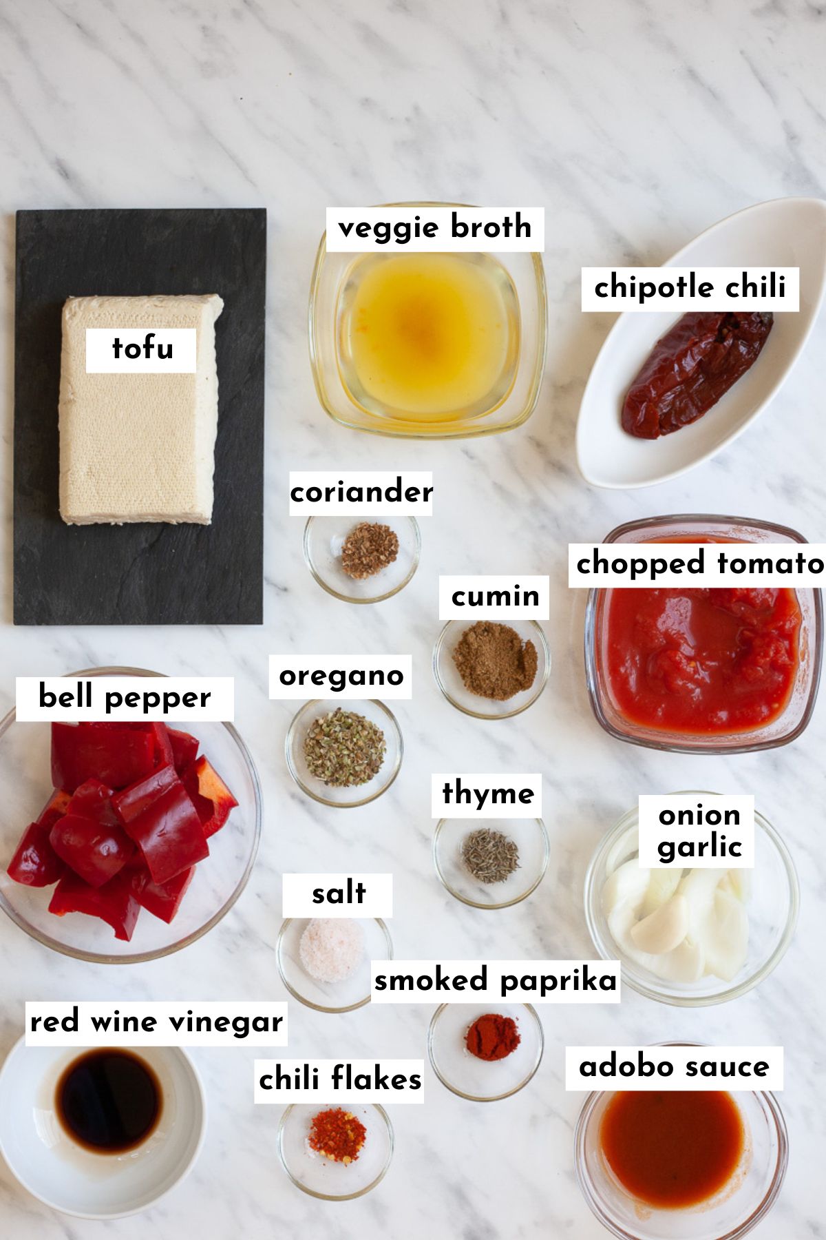 The ingredients of chipotle sofritas in small glass bowls including tofu, veggie broth, chili peppers, chopped red pepper, chopped tomatoes, red sauces and lots of spices. 