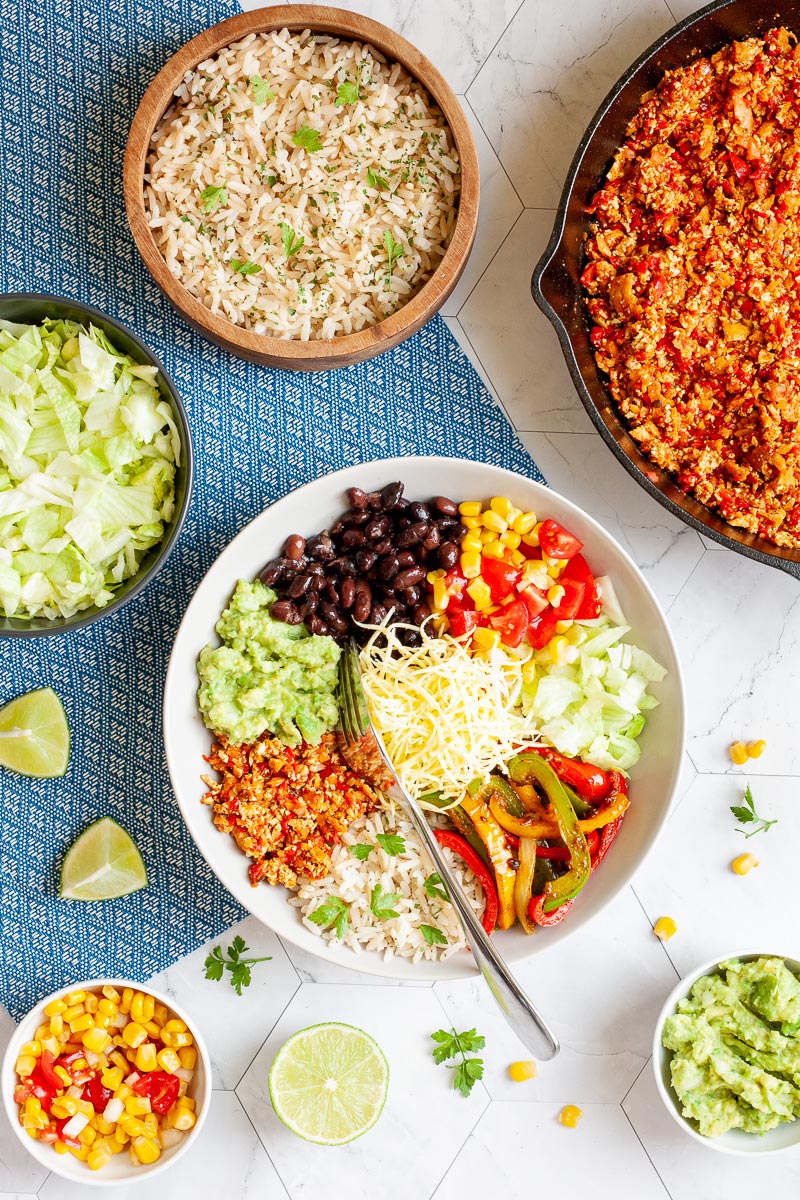 A large white plate in the middle with colorful ingredients like black beans, corn, shredded cheese, bell pepper strips, rice, shredded lettuce, avocado. Small bowls are scattered around with the same ingredients as well as a cast iron skillet with tofu crumbles in red chunky sauce