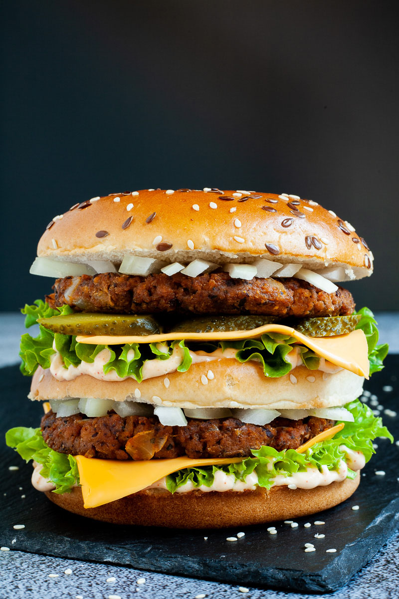 A burger with layers of green ruffled lettuce, yellow sauce, thin cheese slices, dark brown burger patty twice, sliced pickles and white chopped onion.