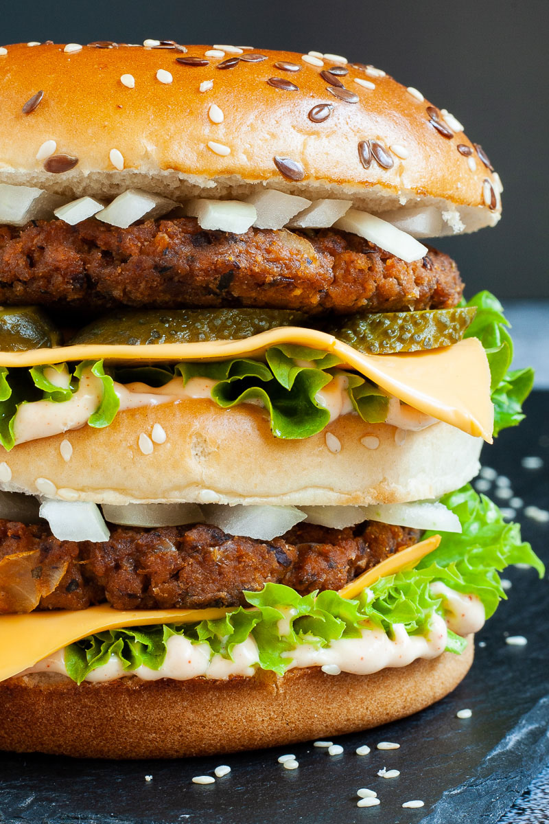 A burger with layers of green ruffled lettuce, yellow sauce, thin cheese slices, dark brown burger patty twice, sliced pickles and white chopped onion.