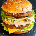 A burger with layers of green ruffled lettuce, yellow sauce, thin cheese slices, dark brown burger patty twice, sliced pickles and white chopped onion.
