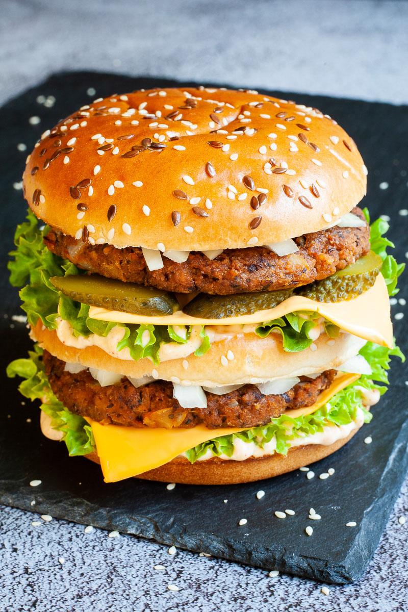 A burger with layers of green ruffled lettuce, yellow sauce, thin cheese slices, dark brown burger patty twice, sliced pickles and white chopped onion.