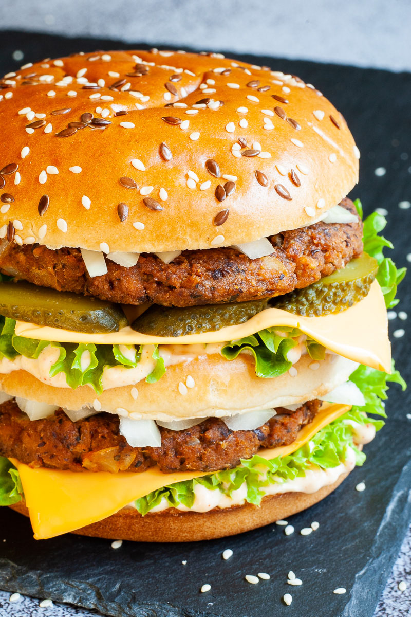 A burger with layers of green ruffled lettuce, yellow sauce, thin cheese slices, dark brown burger patty twice, sliced pickles and white chopped onion.