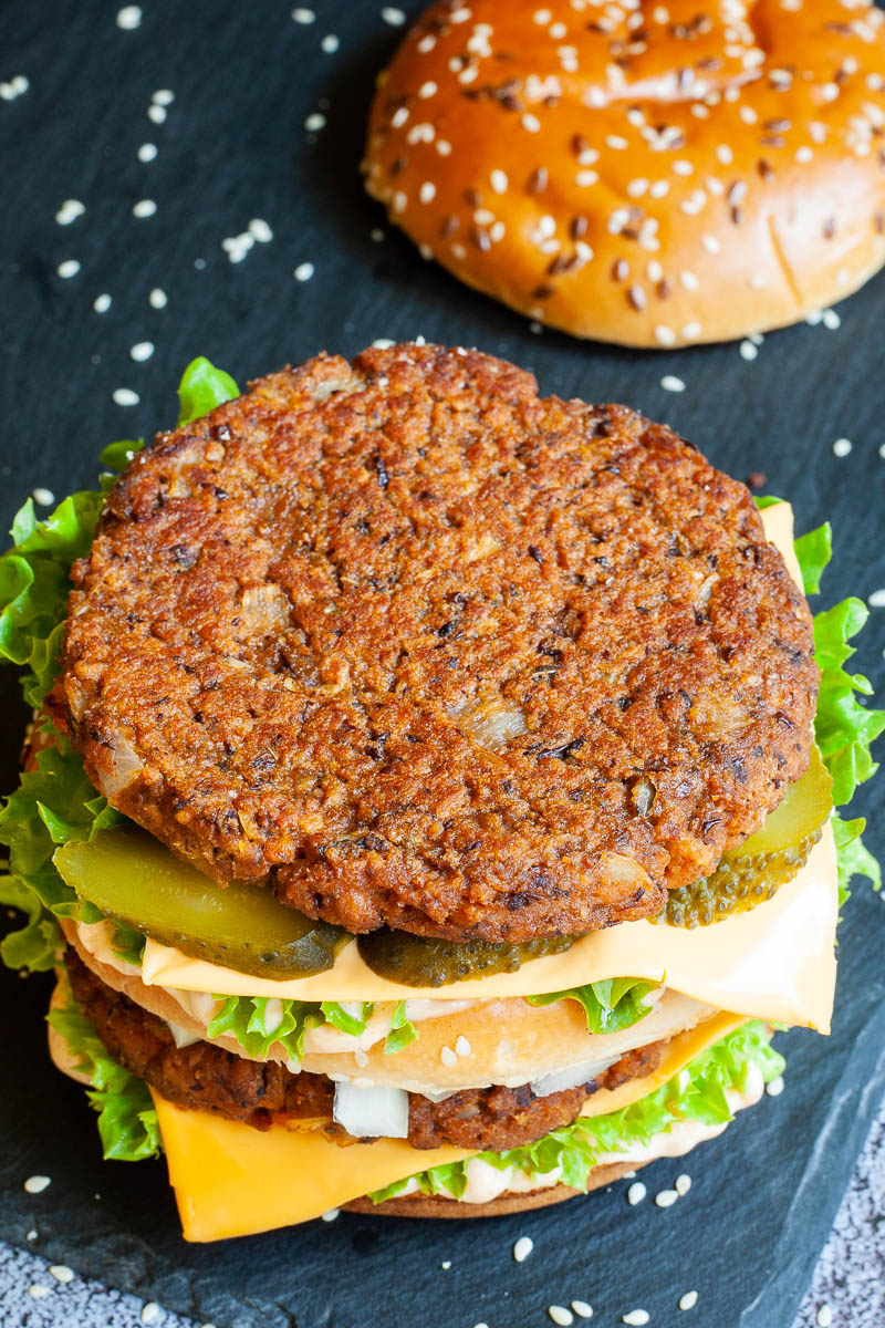 A burger with layers of green ruffled lettuce, yellow sauce, thin cheese slices, dark brown burger patty twice, sliced pickles and white chopped onion. The top bun is right next to it.