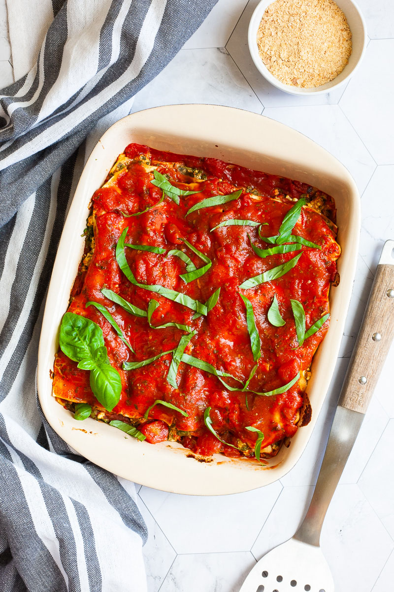 A white plate with a slice of lasagna where you can see the layers of red sauce, pasta sheets and a white ricotto and chopped spinach leaves.