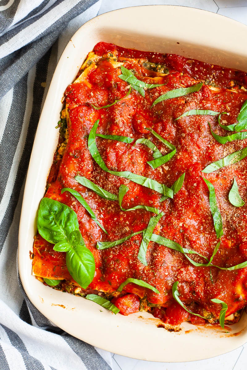 A white plate with a slice of lasagna where you can see the layers of red sauce, pasta sheets and a white ricotto and chopped spinach leaves.