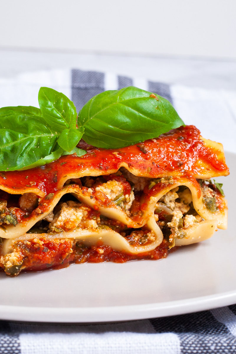 A white plate with a slice of lasagna where you can see the layers of red sauce, pasta sheets and a white ricotto and chopped spinach leaves.
