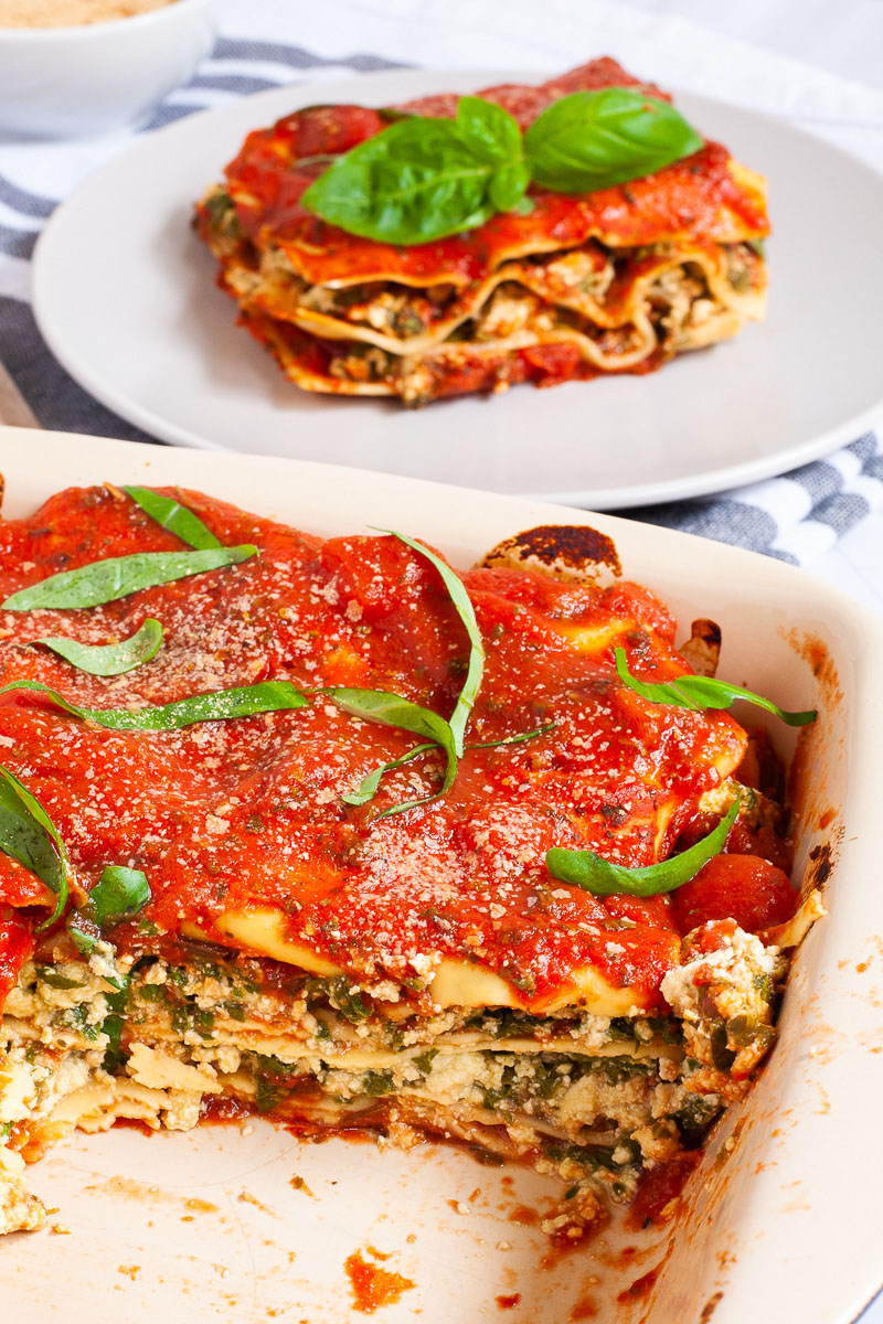 A white plate with a slice of lasagna where you can see the layers of red sauce, pasta sheets and a white ricotto and chopped spinach leaves.