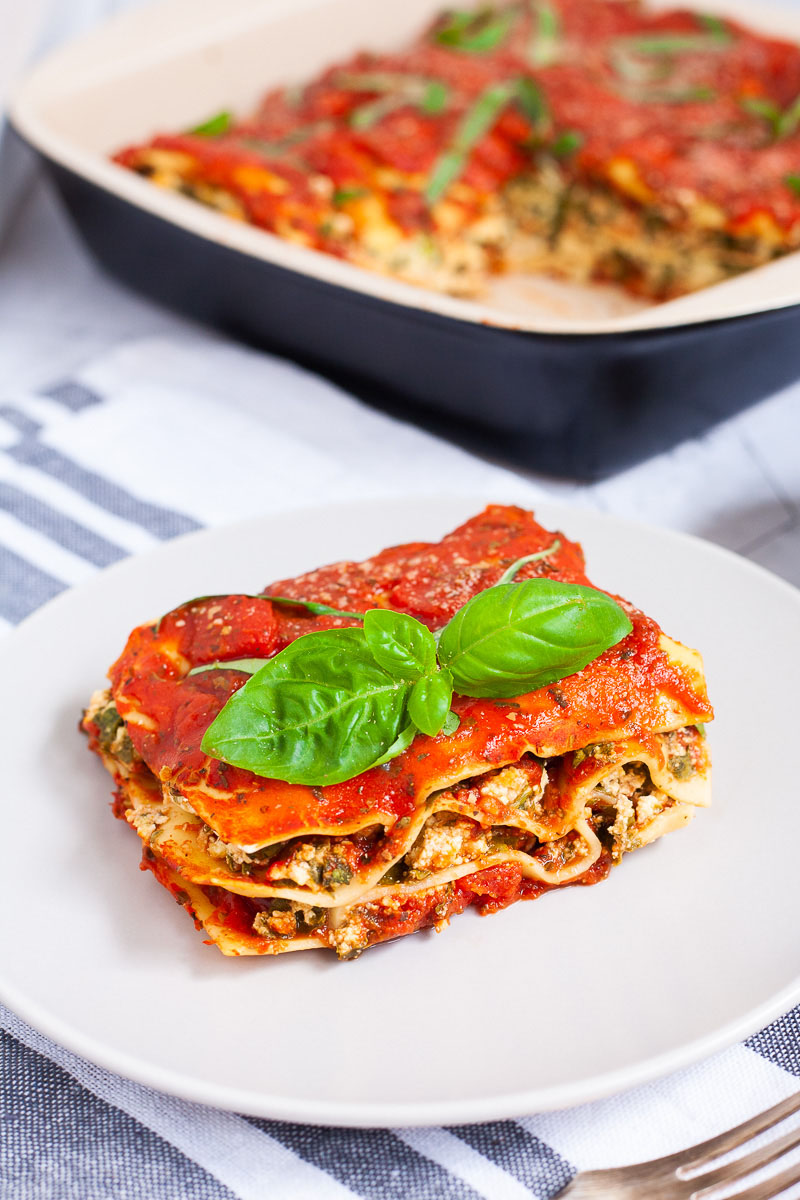 A white plate with a slice of lasagna where you can see the layers of red sauce, pasta sheets and a white ricotto and chopped spinach leaves.