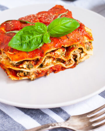A white plate with a slice of lasagna where you can see the layers of red sauce, pasta sheets and a white ricotto and chopped spinach leaves.