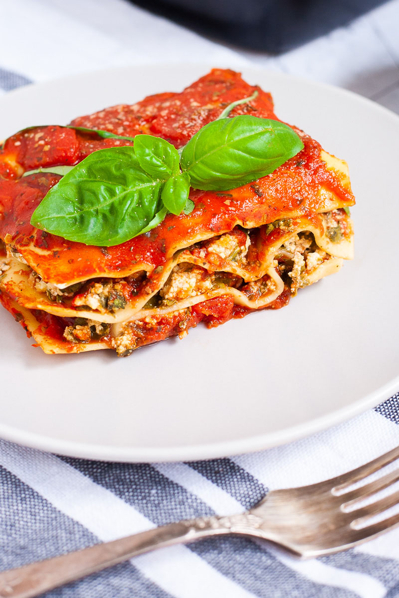 A white plate with a slice of lasagna where you can see the layers of red sauce, pasta sheets and a white ricotto and chopped spinach leaves.