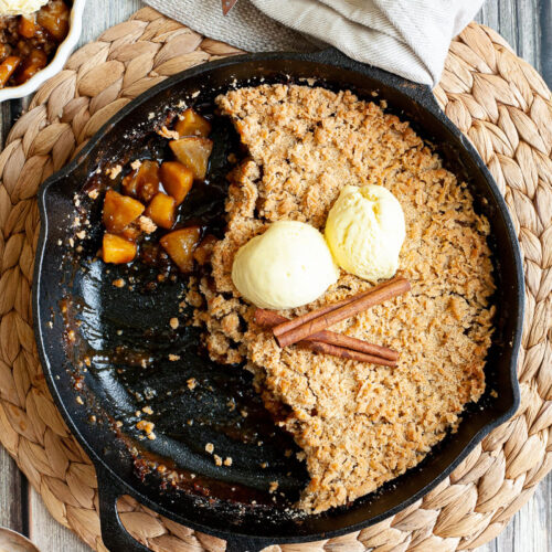 A black skillet from above with diced apples in caramel sauce, crumble topping and 2 scoops of yellow ice cream. 2 small whie bowls is filled with the apple crumble hence there is a part missing in the skillet.