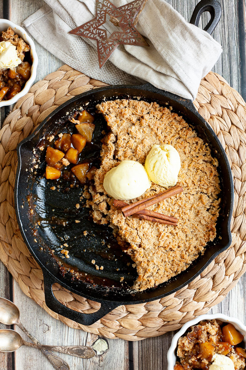 A black skillet from above with diced apples in caramel sauce, crumble topping and 2 scoops of yellow ice cream. 2 small whie bowls is filled with the apple crumble hence there is a part missing in the skillet.