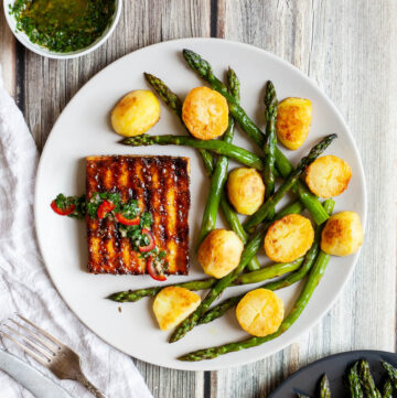 Light grey round plate with a brown, shiny tofu slice topped with a chopped green herbs served with baked half potatoes and roasted asparagus