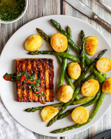 Light grey round plate with a brown, shiny tofu slice topped with a chopped green herbs served with baked half potatoes and roasted asparagus