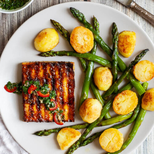Light grey round plate with a brown, shiny tofu slice topped with a chopped green herbs served with baked half potatoes and roasted asparagus