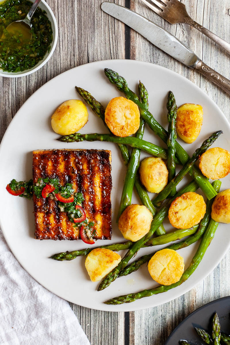 Light grey round plate with a brown, shiny tofu slice topped with a chopped green herbs served with baked half potatoes and roasted asparagus