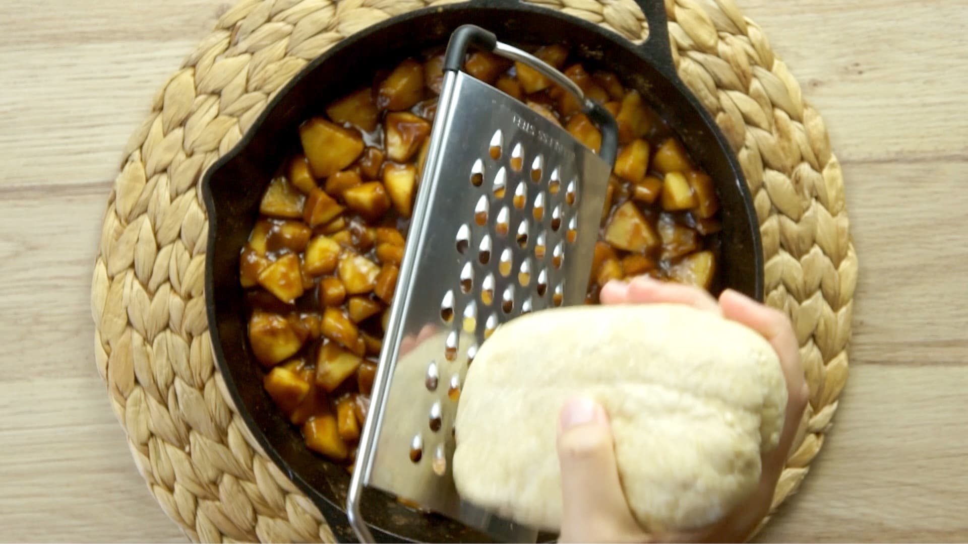 A hand is holding a large hole cheese grater and a frozen dough and about to grate it on top of a black cast iron skillet full of caramel apple slices.