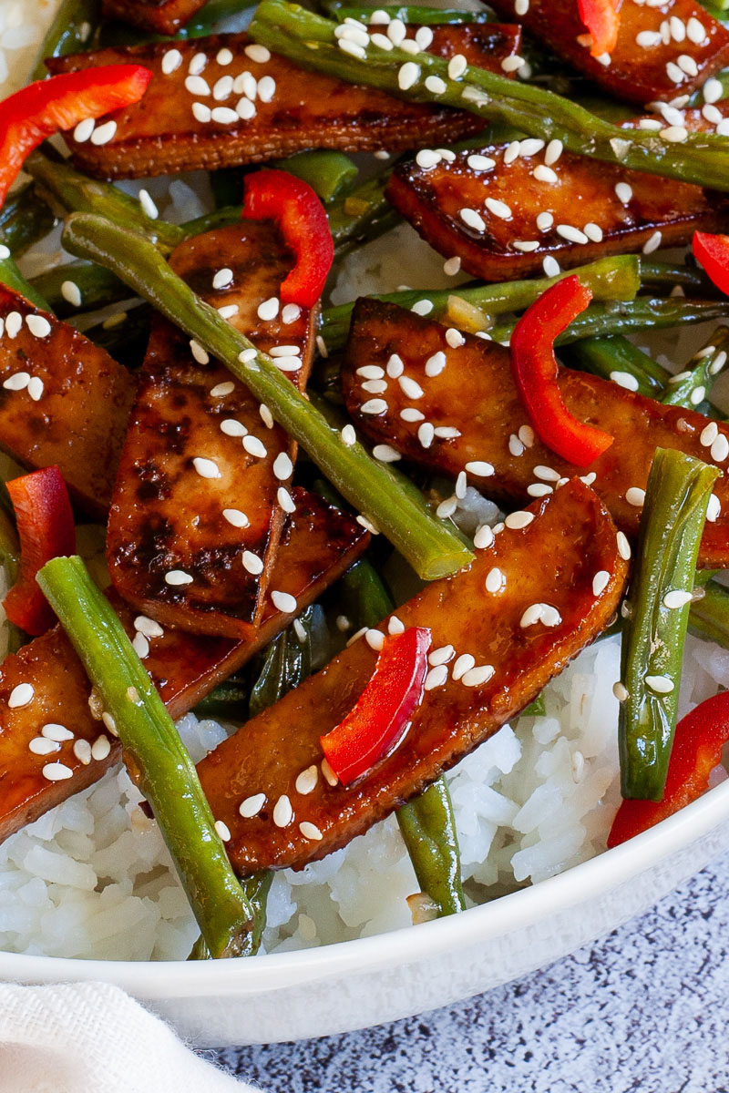White bowl with white rice topped with thin dark brown tofu slices with charred edges, green beans, chopped red bell pepper and sprinkled with white sesame seeds.