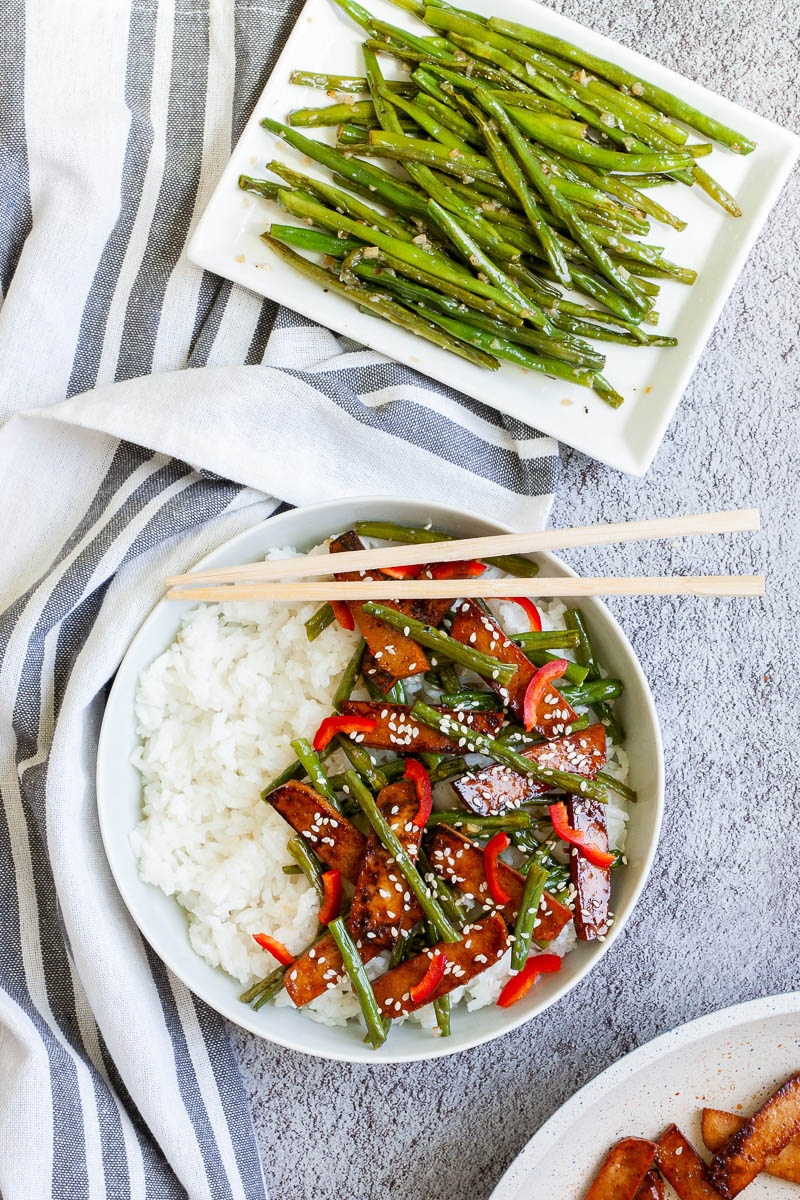 White bowl with white rice topped with thin dark brown tofu slices with charred edges, green beans, chopped red bell pepper and sprinkled with white sesame seeds. Small white plate with shiny sauteed green beans.