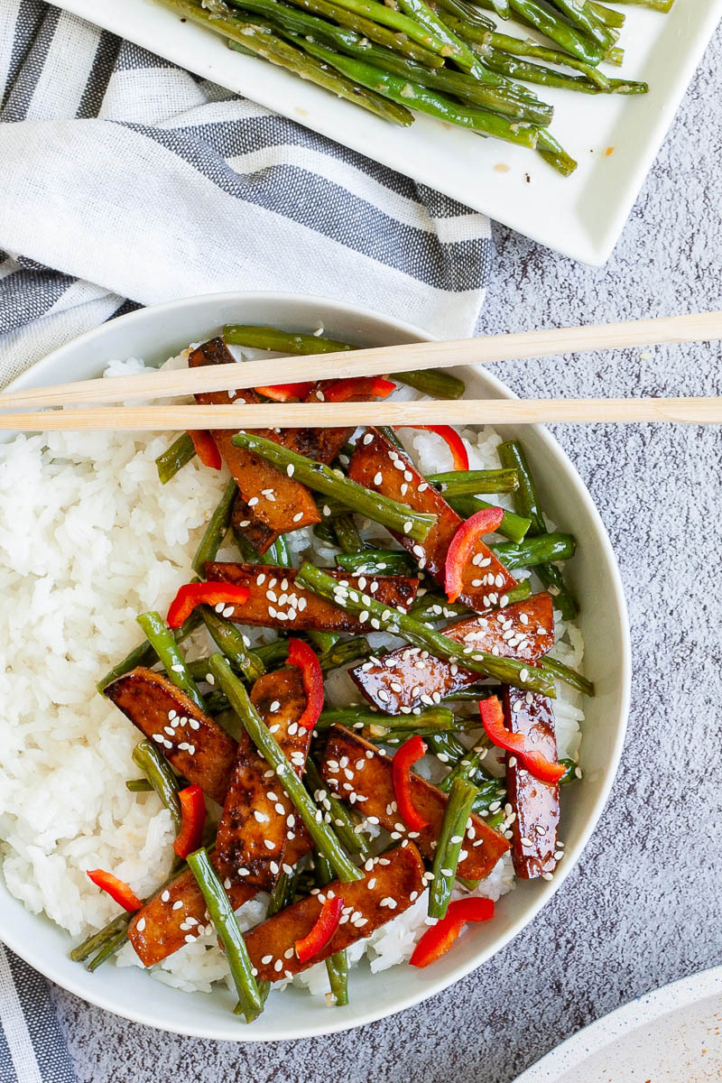 White bowl from above with white rice topped with thin dark brown tofu slices with charred edges, green beans, chopped red bell pepper and sprinkled with white sesame seeds. Light brown chopsticks are placed on the side. Small white plate with shiny sauteed green beans