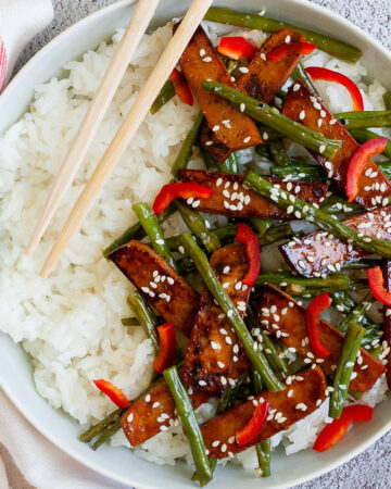 White bowl from above with white rice topped with thin dark brown tofu slices with charred edges, green beans, chopped red bell pepper and sprinkled with white sesame seeds. Light brown chopsticks are placed on the side.